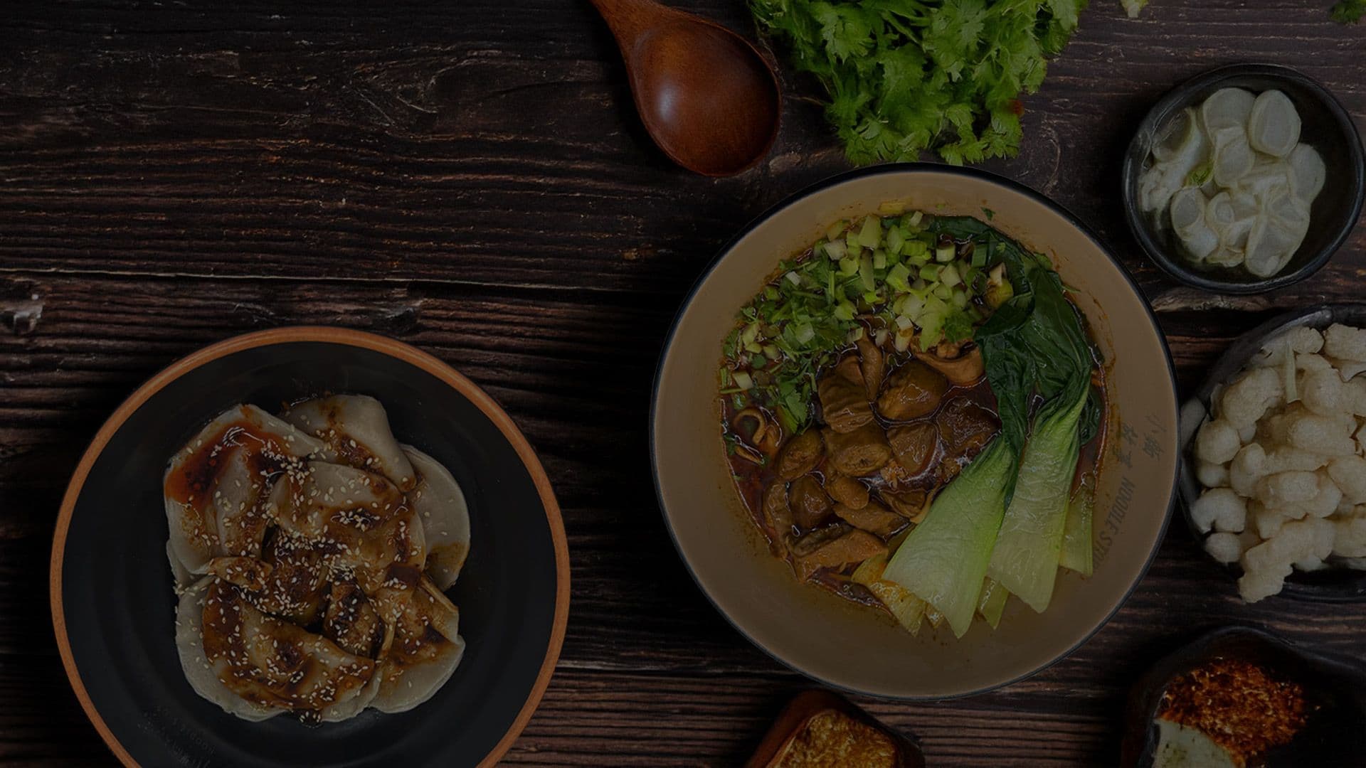 Pan-fried dumplings in chile oil beside a bowl of braised beef noodle soup on a dark wooden table