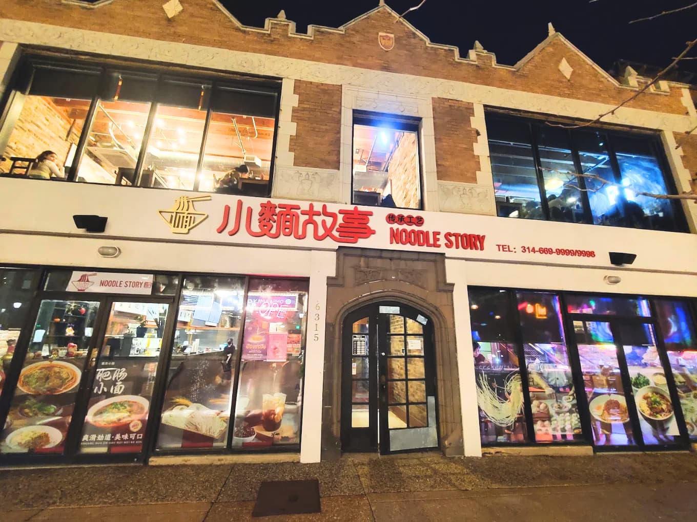 The Noodle Story storefront at night: the lit red 小麵故事 · Noodle Story sign across the brick facade, the arched entrance at 6315, and food posters glowing in the windows.